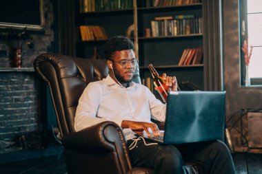 Genç ve mutlu siyah Afrikalı Amerikalı adam oturma odasındaki koltukta dizüstü bilgisayarla iletişim kuruyor.