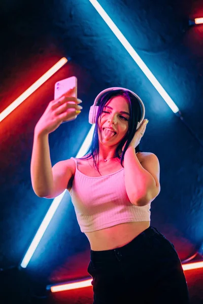 De mooie vrouw mode met koptelefoon luisteren naar muziek en het maken van de selfie foto over rode neon achtergrond in de studio. — Stockfoto