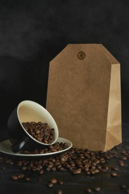 Siyah ahşap masanın üzerinde bir grup kahve çekirdeği ile birlikte sahte el işi kese kağıdı. Siyah dumanlı kahve çekirdekleri.. 