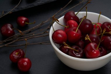 Beyaz bir seramik kasede bir grup kırmızı ve olgun kiraz. Arkaplan olarak ağacın kuru dalları ile siyah bir levha gibi.