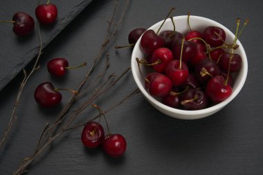 Beyaz bir seramik kasede bir grup kırmızı ve olgun kiraz. Arkaplan olarak ağacın kuru dalları ile siyah bir levha gibi.