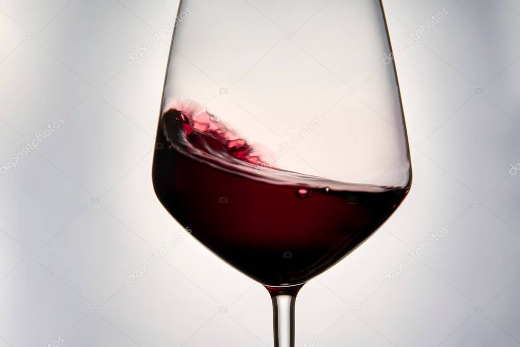 Still-life photography on a white background of a clear glass cup with red wine. The wine creates waves, drops and bubbles inside the glass and is ready to celebrate any event.