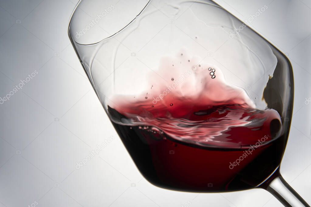 Still-life photography on a white background of a clear glass cup with red wine. The wine creates waves, drops and bubbles inside the glass and is ready to celebrate any event.