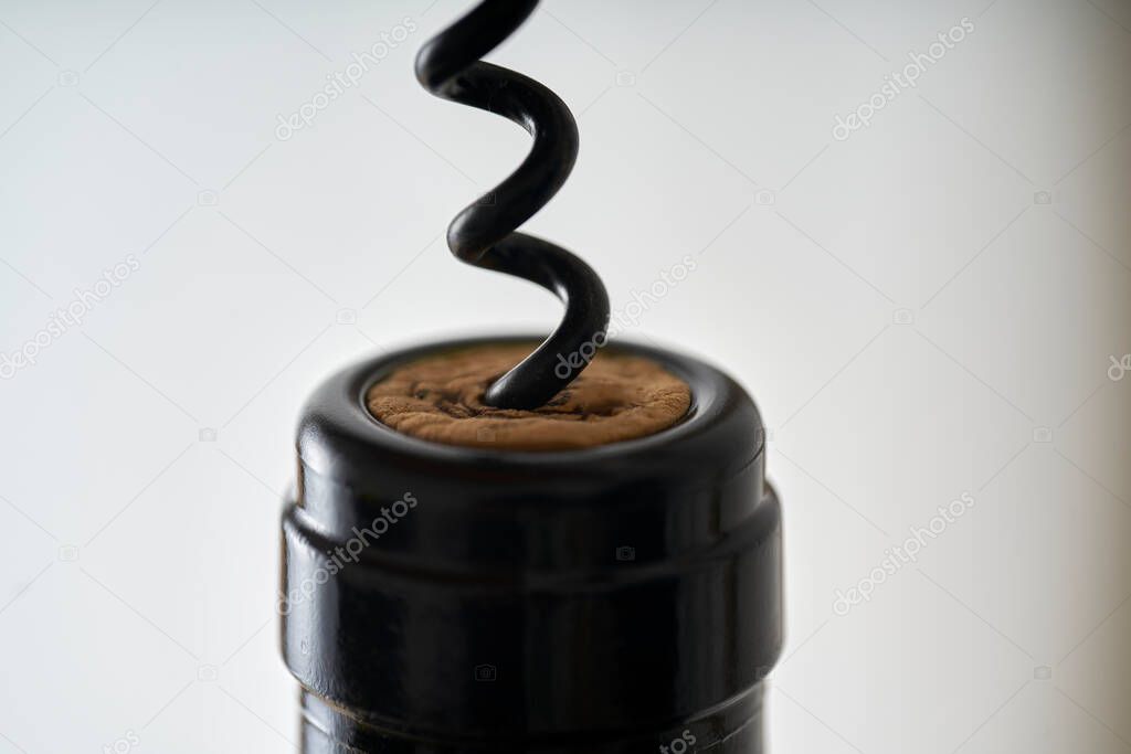 Close-up of a corkscrew opening a bottle of wine. The cork is pierced by the black metal spiral of the tool, which tries to extract it from the dark bottle on a white background.