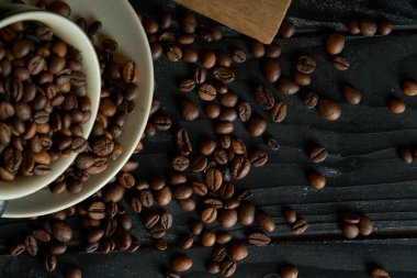 Kavrulmuş siyah kahve çekirdekleri ile dolu seramik fincan, siyah tahta tahtalarda kağıt torba ile fotoğraflanmış ve koyu kahve çekirdekleri.