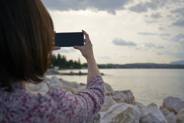Kadının elleri akıllı telefon tutuyor ve gün batımında gölde fotoğraf çekiyor. Seyahat fotoğrafçılığı Garda Gölü 'nün bir anısını çekmiş..