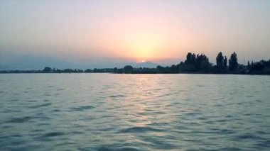 Gölde şafak sökerken ağaçların arasından yükselen güneş. Garda gölü suyu güneşin sıcak renklerini yansıtır, gökyüzü bulutsuzdur ve her şey....