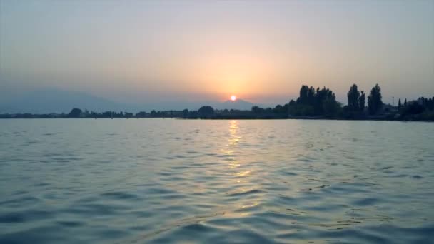 木々の間から山の上に昇る太陽と湖の上で夜明け ガルダ湖の水は 太陽の暖かい色を反映しています 空は雲がなく すべてはかなりあります — ストック動画