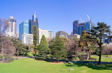 Melbourne 'daki Carlton Gardens' ın huzurlu manzarası, yemyeşil çimenler, olgun ağaçlar ve dekoratif bir gölet. Sahne, modern şehir gökdelenleri ve inşaat vinçlerinin açık mavi gökyüzünün altında bir zemin üzerine kurulmuştur..