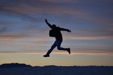 Genç enerjik, mutlu bir adam dağ arka planında yükseğe zıplıyor ve renkli günbatımı bulutları deseni