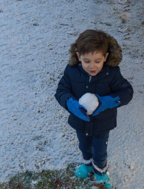 Soğuk bir kış gününde mavi çizme ve büyük bir kartopuyla eldiven giyen çocuk.