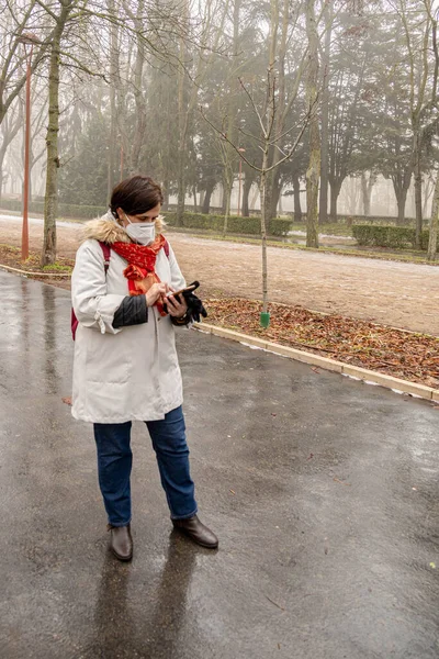 Selektiver Fokus einer warmen Frau mit rotem Rucksack und Gesichtsmaske, die eingehende Nachrichten während eines Stadtspaziergangs mit Kopierplatz per Handy überprüft — Stockfoto