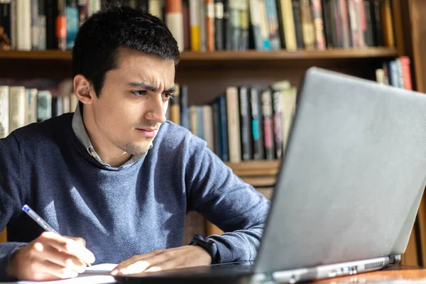 Çalışırken ya da internetten ders çalışırken bilgisayardaki notlara şaşıran ve online çalışma konsepti tablosuna yerleştirilmiş not defterine not alan genç bir adamın seçici odağı..