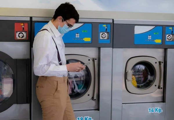 teenager with sanitary mask using cell phone while waiting for laundry to finish cleaning in public washing machine