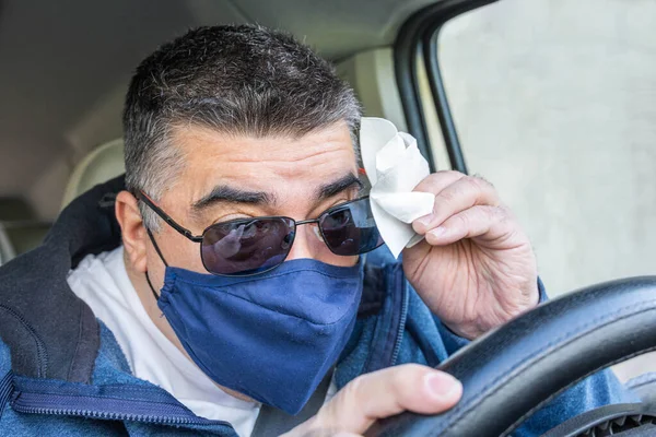 Maskeli bir adam, araba sürerken ve sisli gözlüklerini mendille siliyor çünkü araç konsepti yol güvenliği maskesi kullanırken göremiyor.