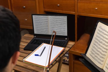 Genç bir adam dizüstü bilgisayarla online çello dersi alıyor ve oturma odasındaki seçici odaklı bir defterin yanındaki yay ile şüpheli notlar işaretliyor..