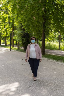 Coronavirus maskeli ve pembe ceketli orta yaşlı bir kadın güneşli bir günde seçici spot ışıkları olan ağaçlarla dolu bir parkta uyum sağlamış bir yolda yürüyor..