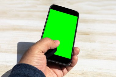 Krom yeşil ekranlı siyah akıllı telefonu kullanarak sanki bir mesaj yolluyor ya da güneşli bir günde açık ahşap bir masaya bir uygulama tıklatıyormuş gibi.