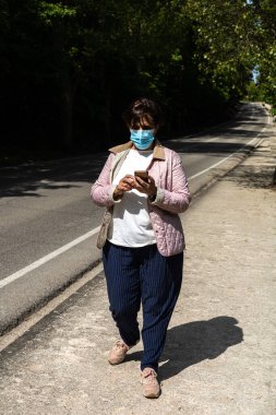 Koronavirüs ya da kirlilik maskesi takan genç bir kadın güneşli bir günde yol kenarında yürürken akıllı telefonuyla mesaj atıyor.