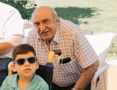 portrait of a grandfather with mustache with his grandson with sunglasses and disability in a wheelchair smiling on a summer's day