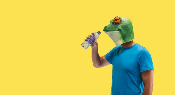 コピースペースで黄色の背景に生態系を害するペットボトルから新鮮な飲料水を飲むカエルマスクを持つ人のジェスチャー — ストック写真