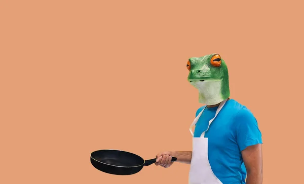 Kurbağa şef maskeli, önlüklü bir insan jesti ve portakal arkaplanda fotokopi alanı ile yemek pişirmek ya da öğrenmek için bir tava tutuyor.