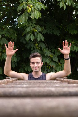 Gülümseyen ve ellerini kaldıran genç sporcu korkuluk jimnastik egzersizleri yapmak için tahta parmaklıkların üzerinde.