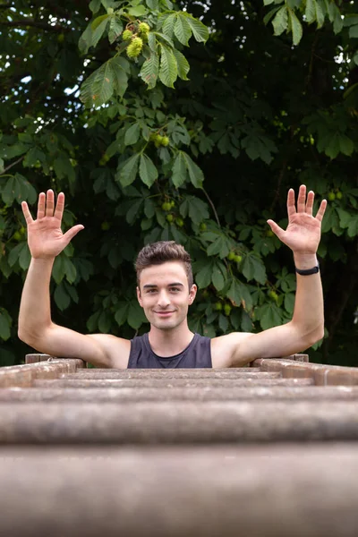Gülümseyen ve ellerini kaldıran genç sporcu korkuluk jimnastik egzersizleri yapmak için tahta parmaklıkların üzerinde.