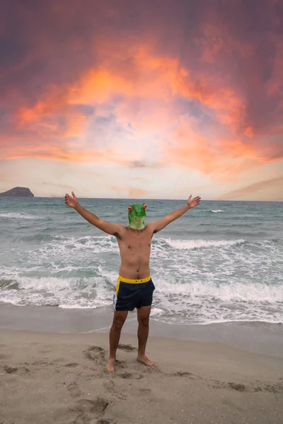 Jonge man met badpak en kikkermasker die zijn armen uitstrekt aan de kust van het strand met een roodachtige lucht — Stockfoto