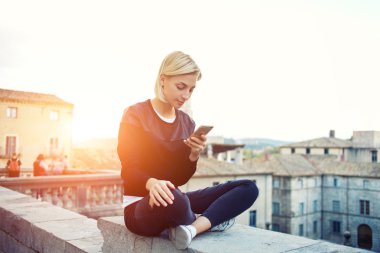  Hipster kız cep telefonumetinden kısa mesaj okuyor, bir binanın çatısında otururken