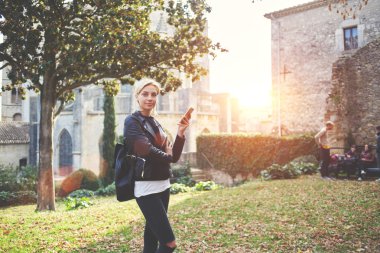 Young stylish female is holding cell telephone, while is standing outside