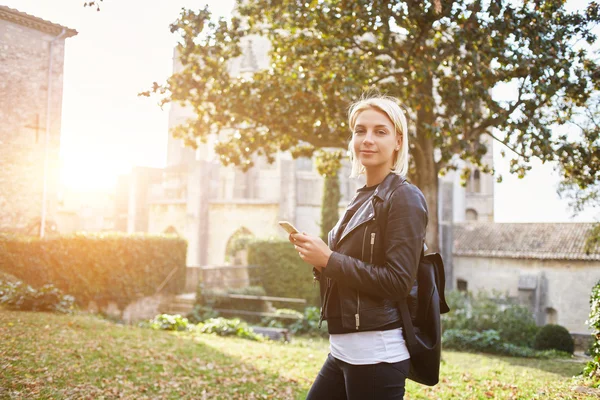 Elegante chica hipster está posando con el teléfono celular en las manos, mientras está de pie al aire libre en el día —  Fotos de Stock