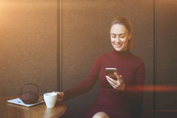 Fiatal, mosolygós nő olvasó szöveges üzeneteket a mobiltelefon, míg pihen a kávézó belső — Stock Fotó
