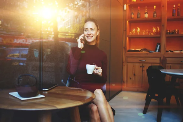Χαρούμενα θηλυκό καλώντας με έξυπνο τηλέφωνο ενώ κάθεται σε μοντέρνο εσωτερικό εστιατόριο — Φωτογραφία Αρχείου