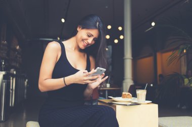 Young hispter girl is watching funny video on cell telephone whille is sitting in coffee shop