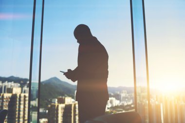Erkek emploee ellerde hücre telefon ile büyük gökdelen penceresinde şehir manzaralı yakınındaki ofiste duruyor