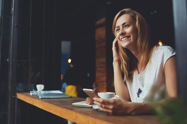 Charming smiling hipster girl received good news on cell telephone while she sitting in cozy coffee shop