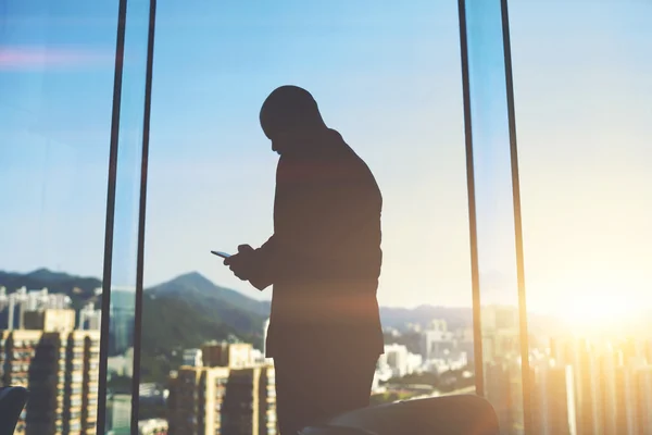 Male emploee with cell telephone in hands is standing in office near big skyscraper window with city view — Stock Photo, Image