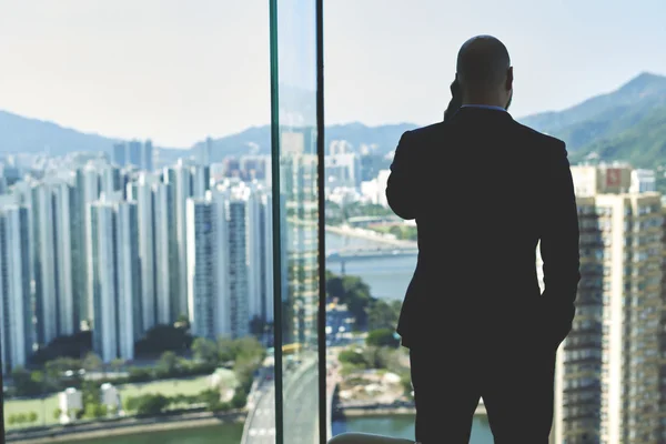 Joven abogado masculino está llamando con teléfono celular, mientras está de pie cerca de la ventana de la oficina con vista de rascacielos altos — Foto de Stock