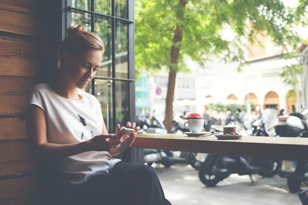 Attraktive Frau liest SMS auf dem Handy, während sie in einem gemütlichen Café sitzt — Stockfoto