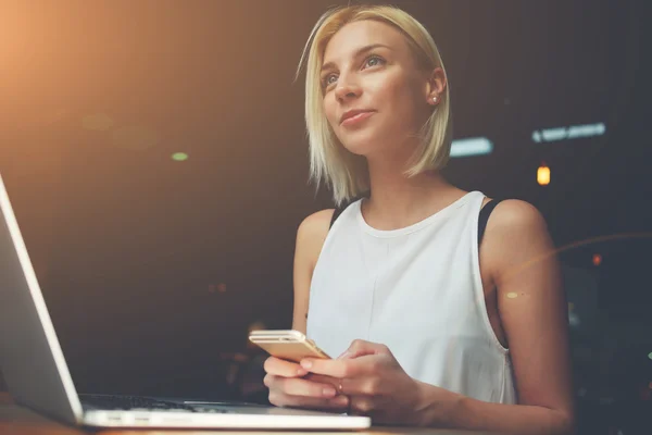 Charmante femme rêveuse utilisant un téléphone cellulaire et un ordinateur portable pendant le repos dans un café — Photo