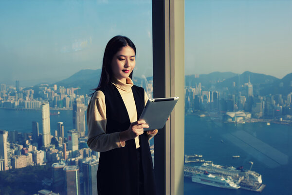 Female is using touch pad, while is standing in modern office interior against window with view developed New York cityscape