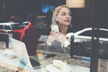 Young woman is using mobile phone, while is sitting in modern cafe-bar with portable laptop computer