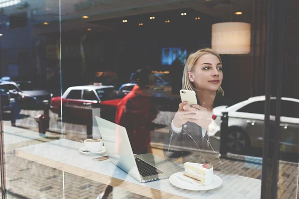 Joven estudiante hermosa mujer está utilizando el teléfono celular, durante el desayuno en la cafetería y el trabajo en el ordenador portátil portátil — Foto de Stock