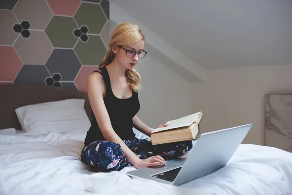Encantadora estudiante de gafas está buscando material de estudio en Internet a través de computadora portátil, mientras está sentada en el interior de su casa —  Fotos de Stock