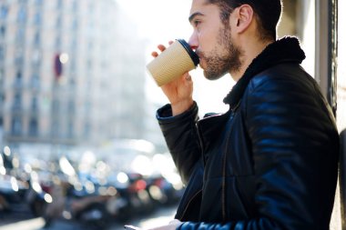 Sıcak siyah ceket giyen sakallı bir erkek güneşli bir günde sokakta dikilirken bir fincan taze kahve içiyor.