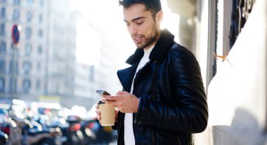 Modern binanın yakınındaki şık kıyafetli, sakallı bir erkek. Elinde kahve fincanı ve cep telefonu var.