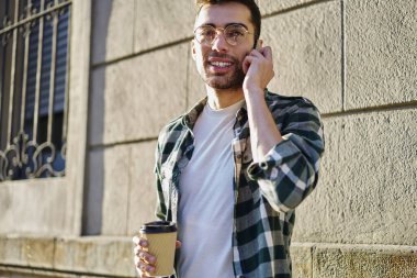 Kareli tişörtlü ve gözlüklü genç, düşük açılı, güneşli bir günde modern binanın duvarında dikilirken gözlüklü, kahve içen ve cep telefonuyla konuşan bir adam.