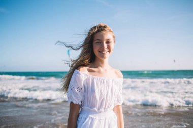 Yazlık elbiseli, pozitif gülümseyen küçük sarışın kumlu sahilde duruyor parlak güneşin altında sallanıyor ve kameraya bakarken gülümsüyor.