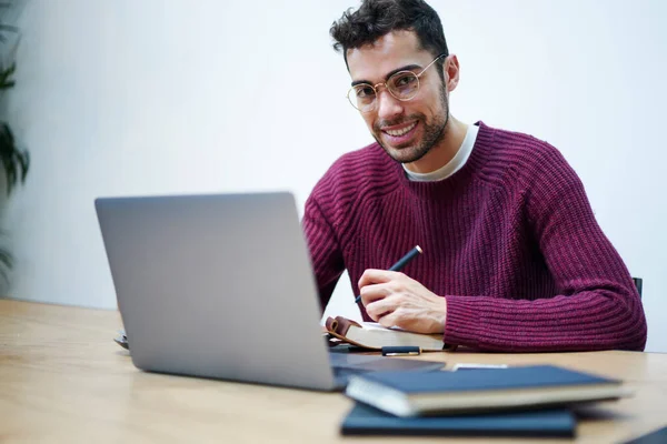 Cheerful ethnic bearded male freelancer in casual wear and glasses sitting at wooden table with laptop and writing thoughts in copybook while looking at camera